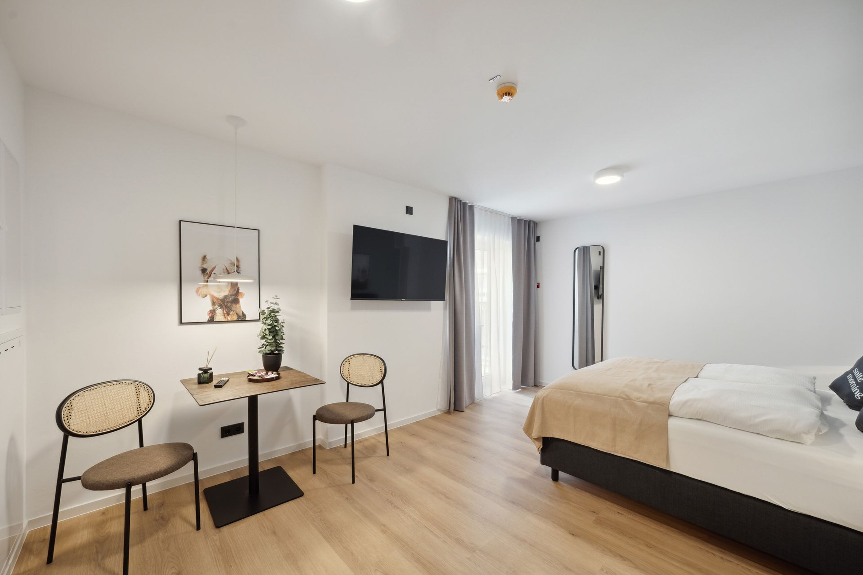 A modern hotel room with a bed, a desk with chairs, a TV, and a framed picture on the wall.
