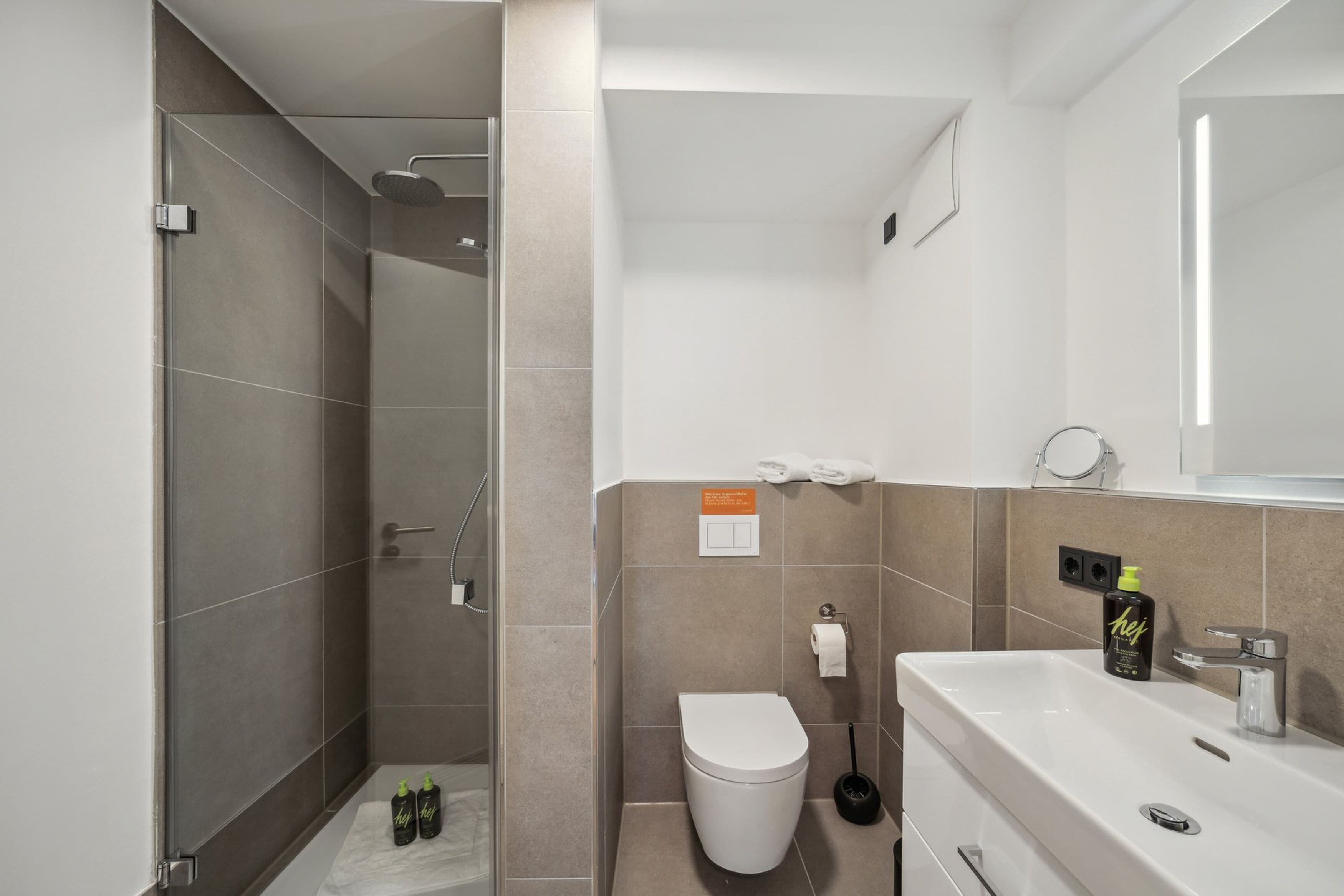 A modern bathroom with a shower, toilet, and sink, featuring a mirror, towel rack, and a sign on the wall.