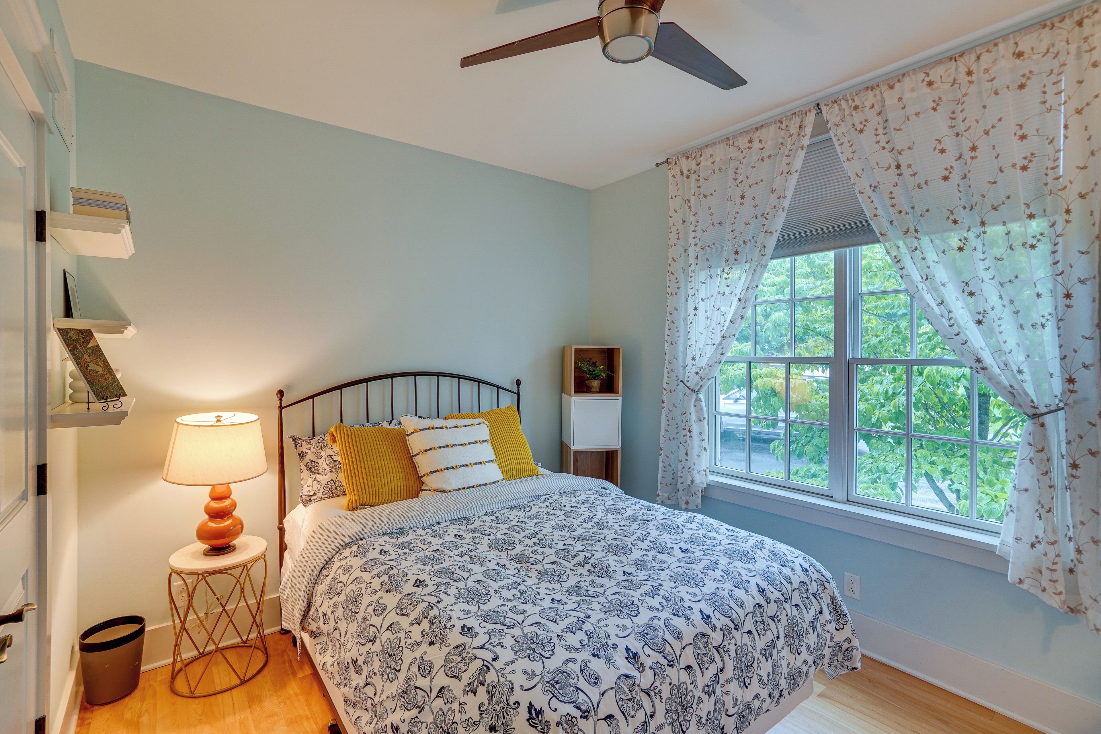 Une chambre à coucher avec un lit, une table de chevet avec une lampe, une fenêtre avec des rideaux et un ventilateur de plafond.