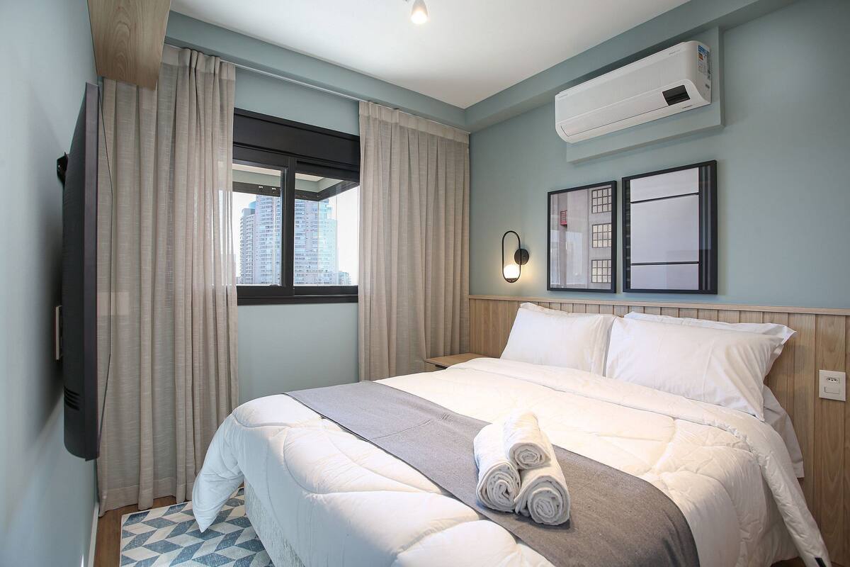 Ein Hotelzimmer mit Bett, Vorhängen, Klimaanlage, Wandleuchte und Fenster mit Blick auf Gebäude.