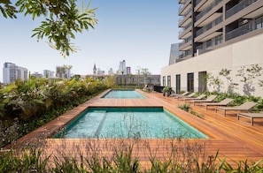 A rooftop pool with wooden decking, lounge chairs, and a city skyline view. - Expand Pinheiros (São Paulo)