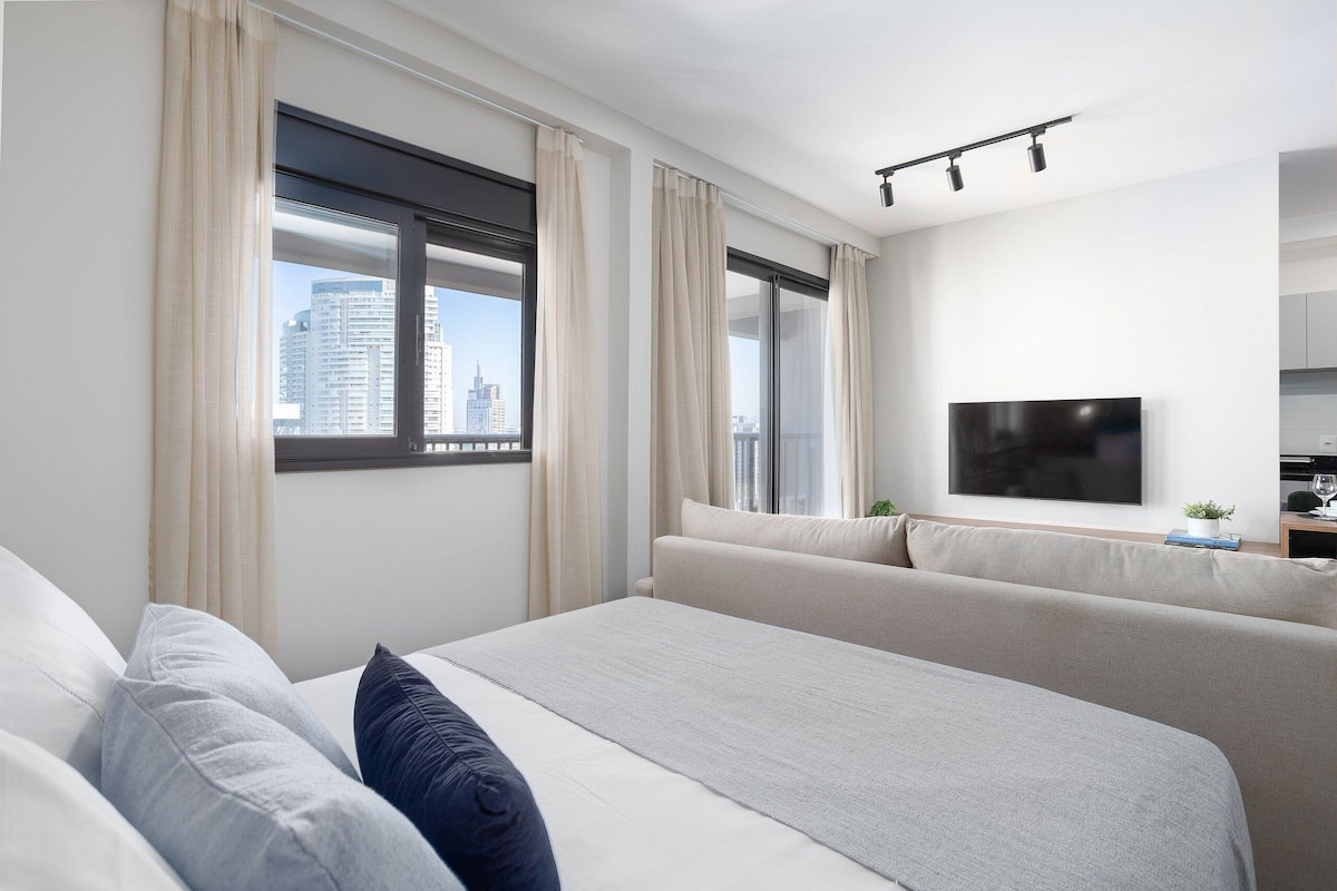 A modern hotel room with a large bed, a sofa, a TV, and a window with curtains.