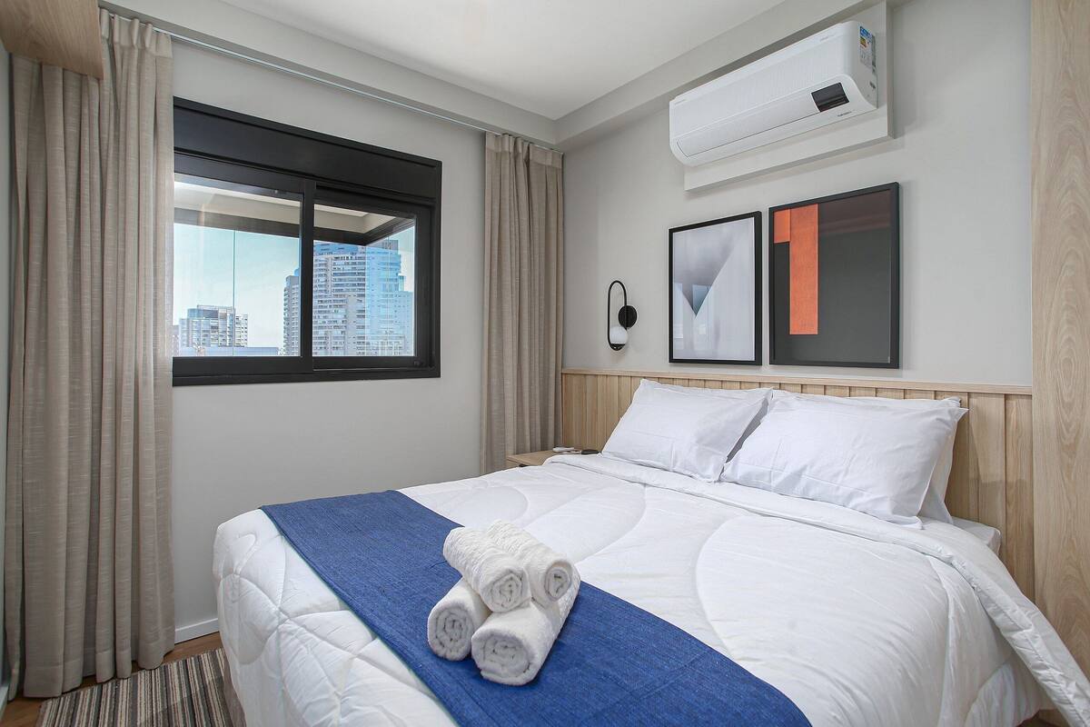 Ein Hotelzimmer mit Bett, einem fensterbepflanzten Fenster, einer Klimaanlage und gerahmten Bildern an der Wand.