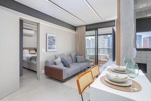 Uma sala de estar moderna com sofá, mesa de jantar e cadeiras, varanda com vista para a cidade, e um quarto separado com cama e janela.