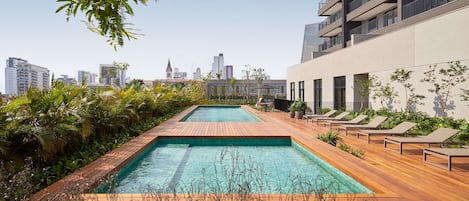 A rooftop pool with wooden decking, lounge chairs, and a city skyline view.