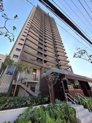 Um edifício alto com várias varandas, entrada com portão e áreas verdes ao redor.
