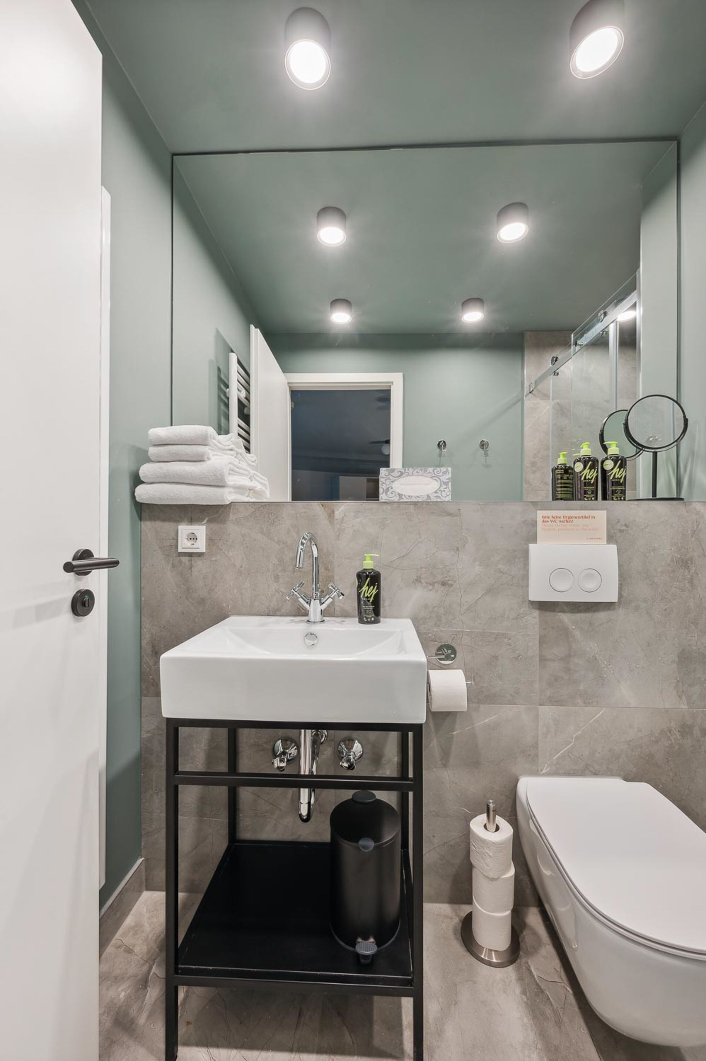 Un baño moderno con un lavamanos blanco, un mueble negro y un inodoro. Hay toallas apiladas en un estante, y un espejo sobre el lavamanos.