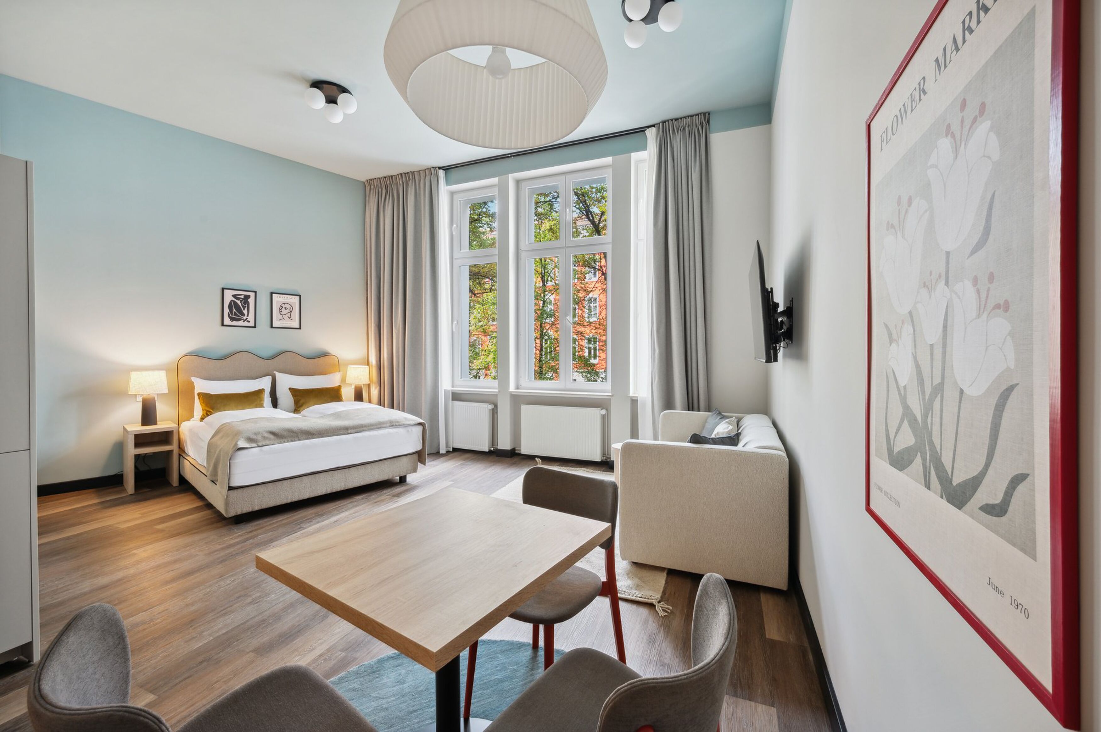 A hotel room with a bed, bedside tables, a desk, chairs, a large window with curtains, and a wall-mounted light fixture.