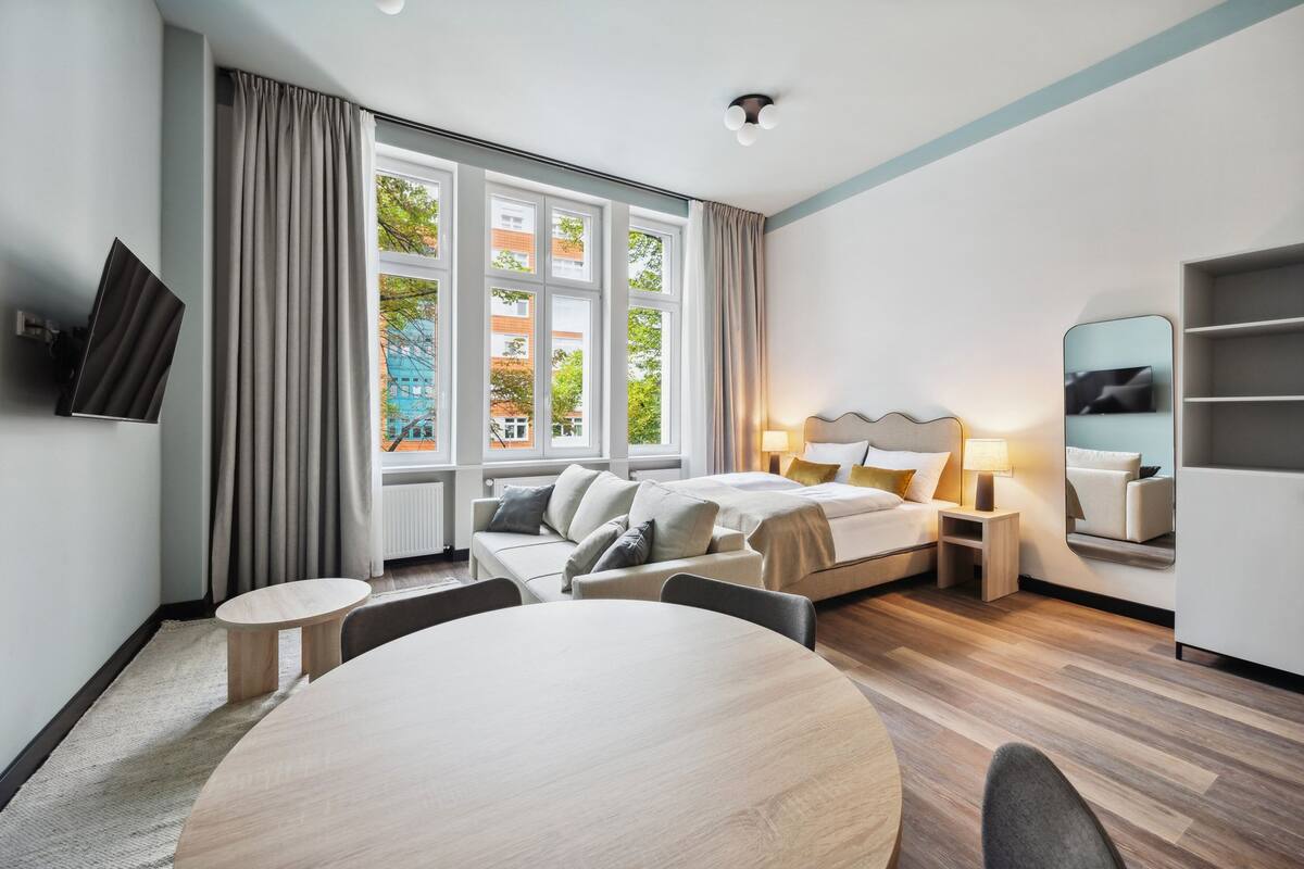 Una habitación de hotel moderna con un ventanal grande, una cama con cabecero, una mesita de noche con lámparas, una mesa redonda y un televisor montado en la pared.