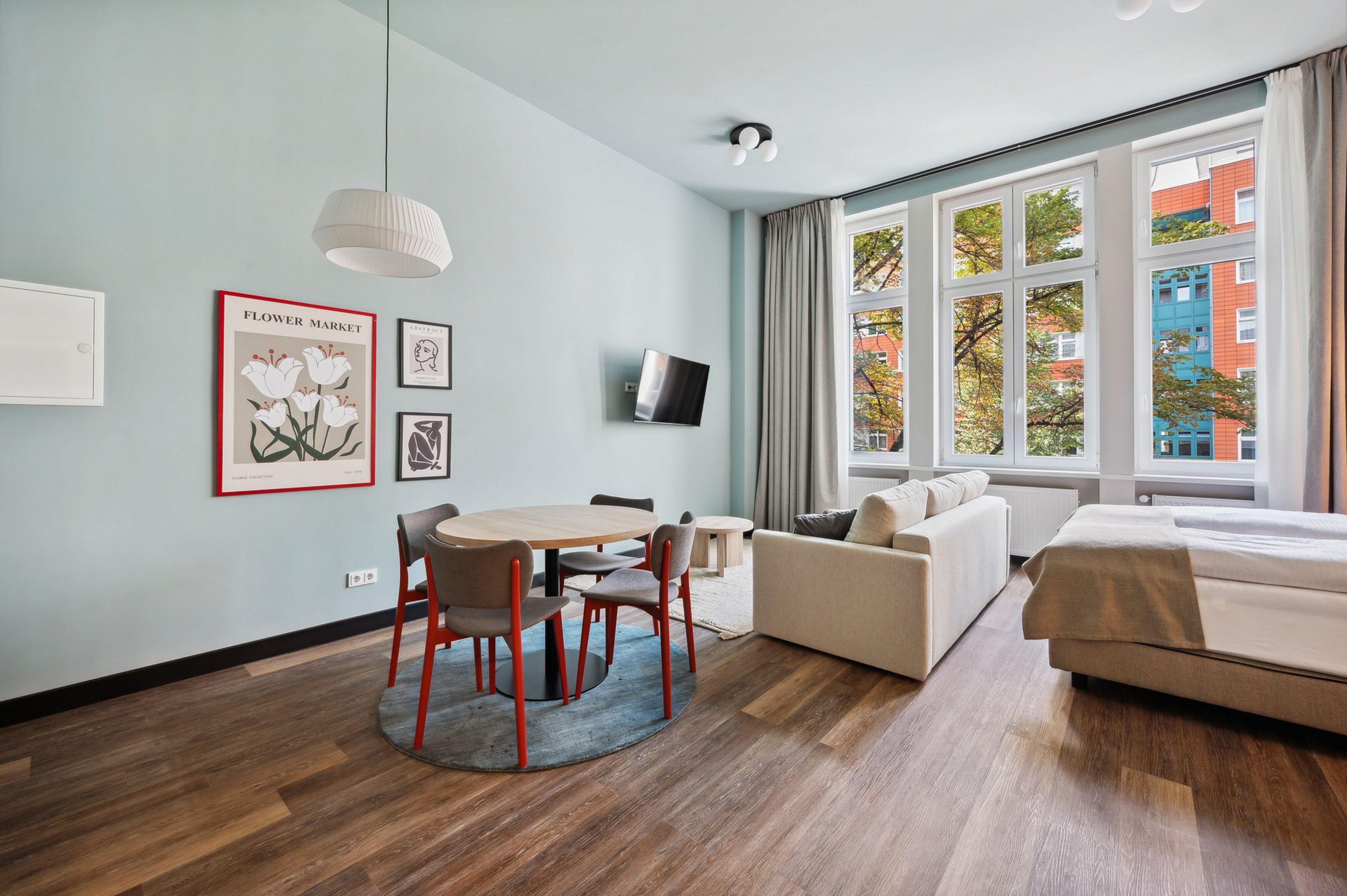 Une chambre d’hôtel moderne avec un plancher en bois, une table de salle à manger ronde entourée de chaises, un canapé, un lit, un téléviseur fixé au mur et de grandes fenêtres offrant une vue sur la ville.