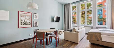 Une chambre d’hôtel moderne avec un plancher en bois, une table de salle à manger ronde entourée de chaises, un canapé, un lit, un téléviseur fixé au mur et de grandes fenêtres offrant une vue sur la ville.