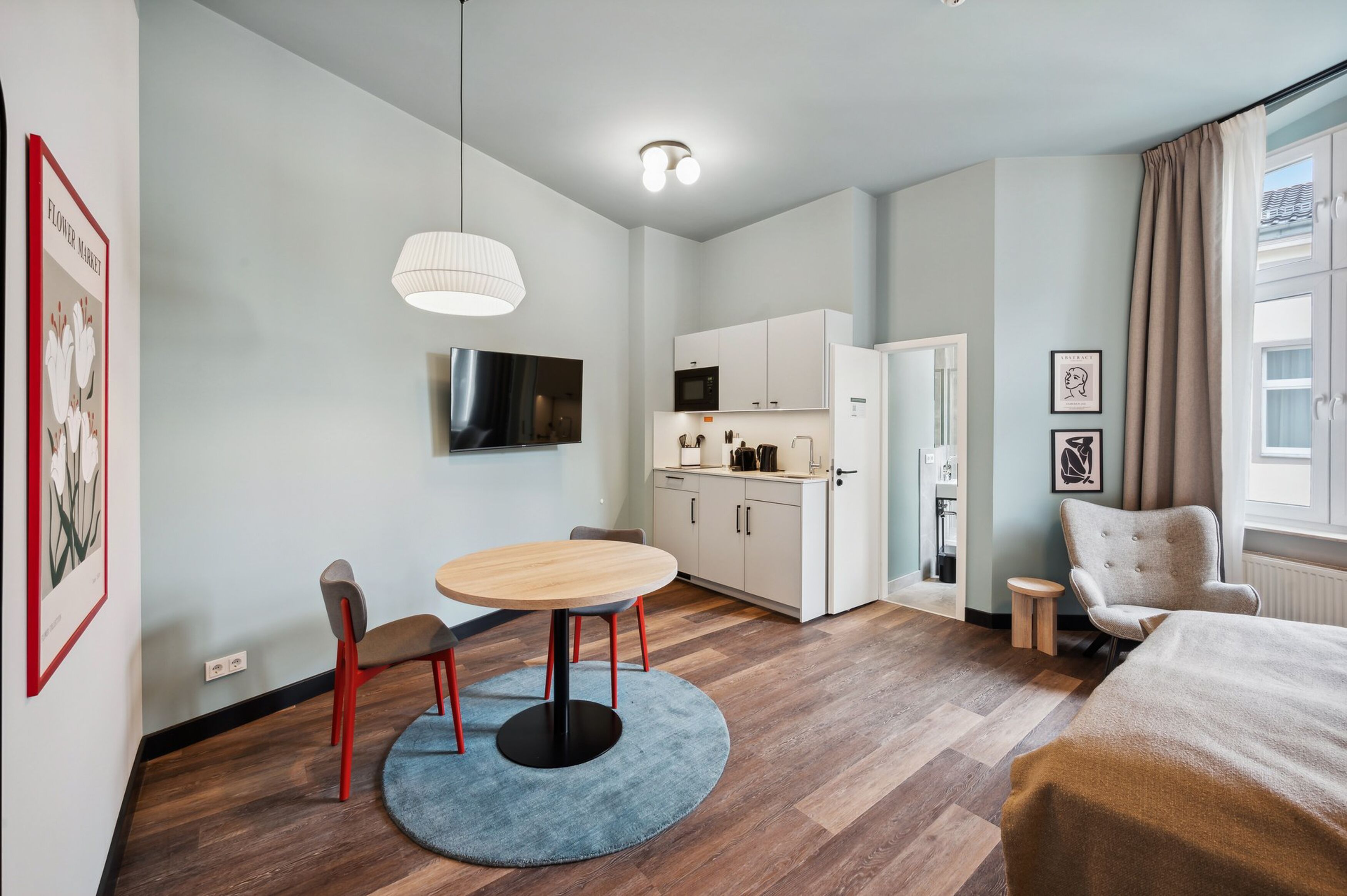 A modern hotel room with a dining area, kitchenette, and a large window with curtains.