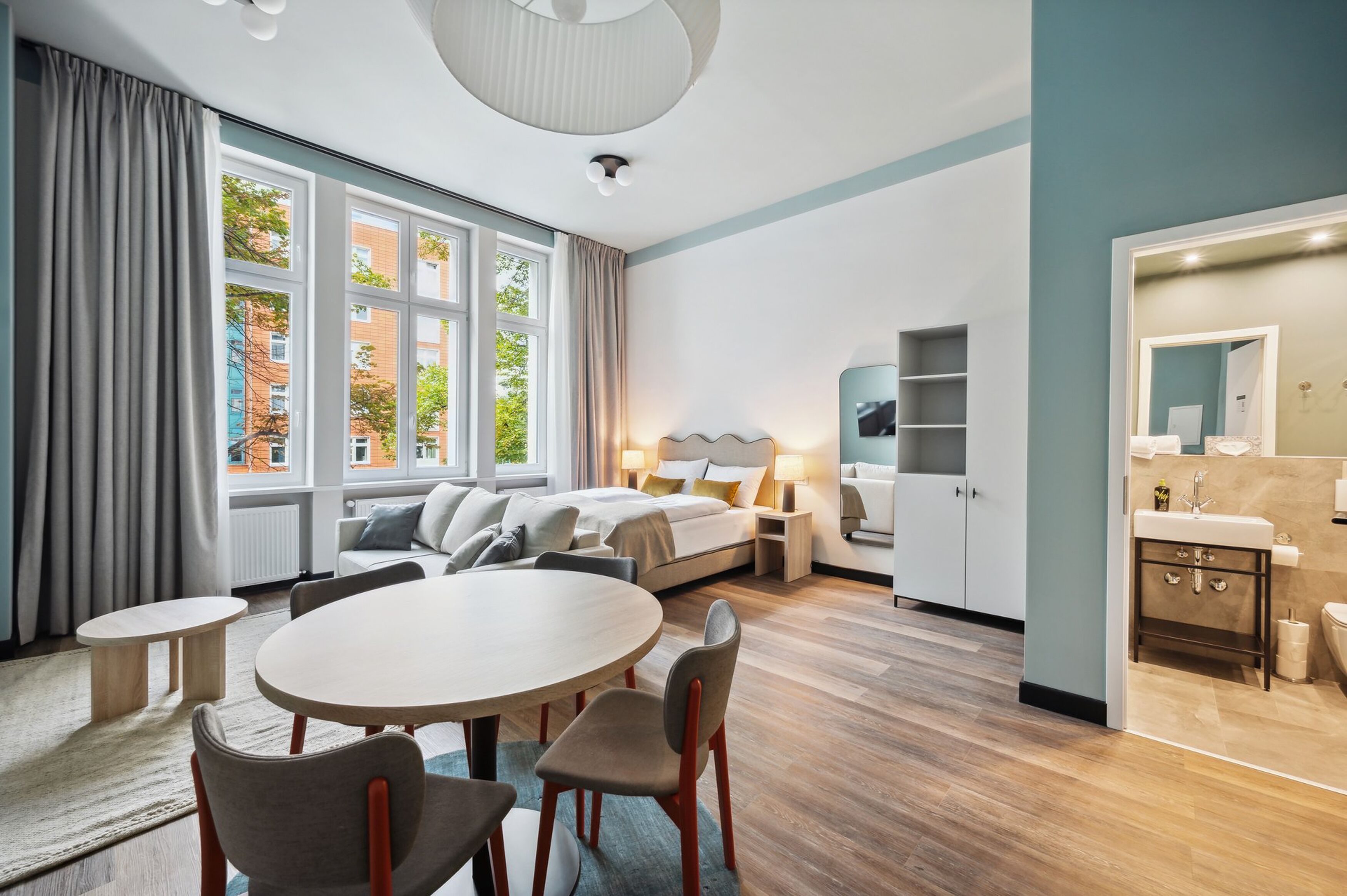 A modern hotel room with a large bed, a dining table, and a bathroom with a mirror and sink.