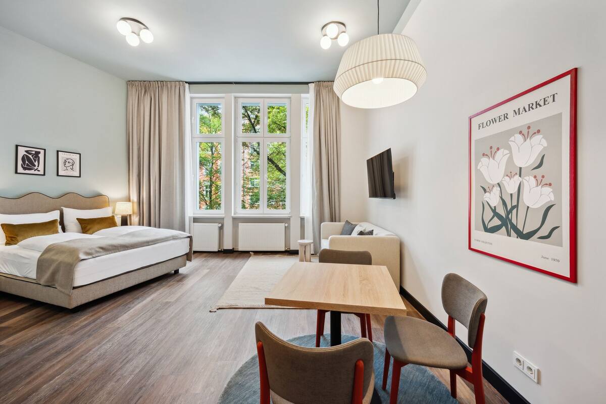 Una habitación de hotel moderna con una cama, una mesa de comedor con sillas, un televisor y un amplio ventanal con cortinas.