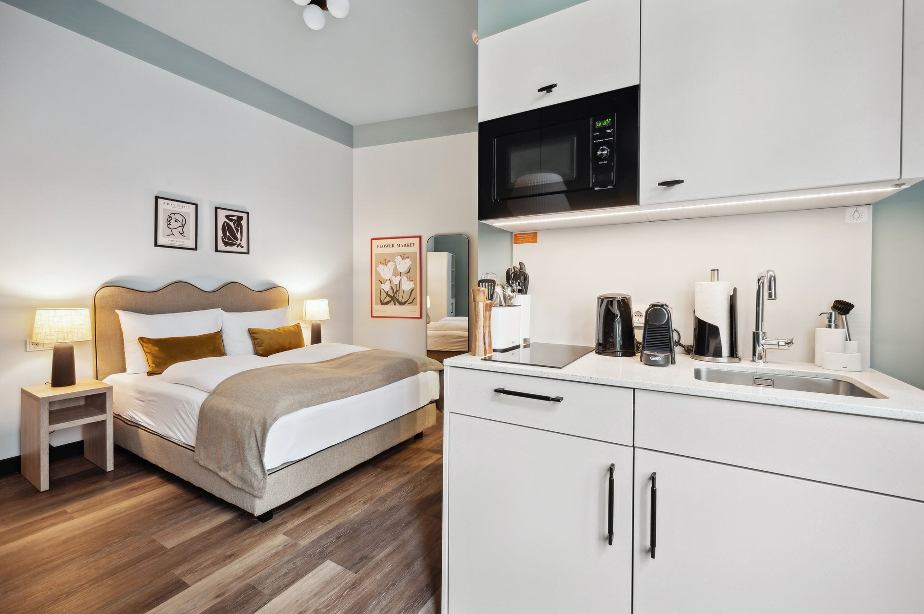 Una habitación de hotel moderna con cama, mesita de noche, lámpara y una kitchenette con microondas, lavamanos y varios electrodomésticos.
