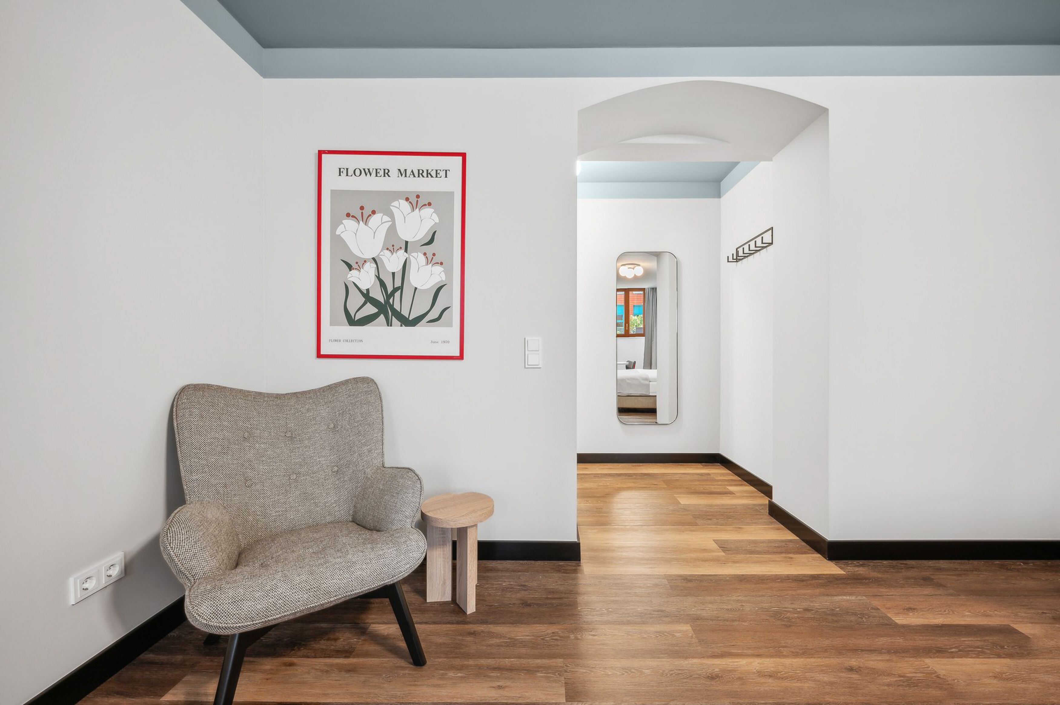 A hallway with a grey upholstered chair, a small wooden side table, and a framed picture of flowers on the wall.