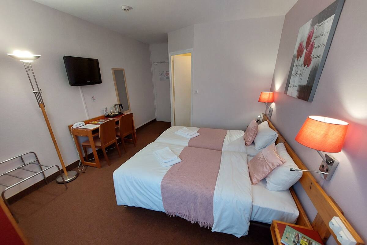 A hotel room with a bed, bedside table, lamp, desk with chair, and a television on the wall.