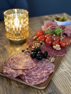 A wooden board with various meats and olives, a lit candle, and a bowl of cherry tomatoes on a wooden table. - LE 120 (La Chataigneraie)