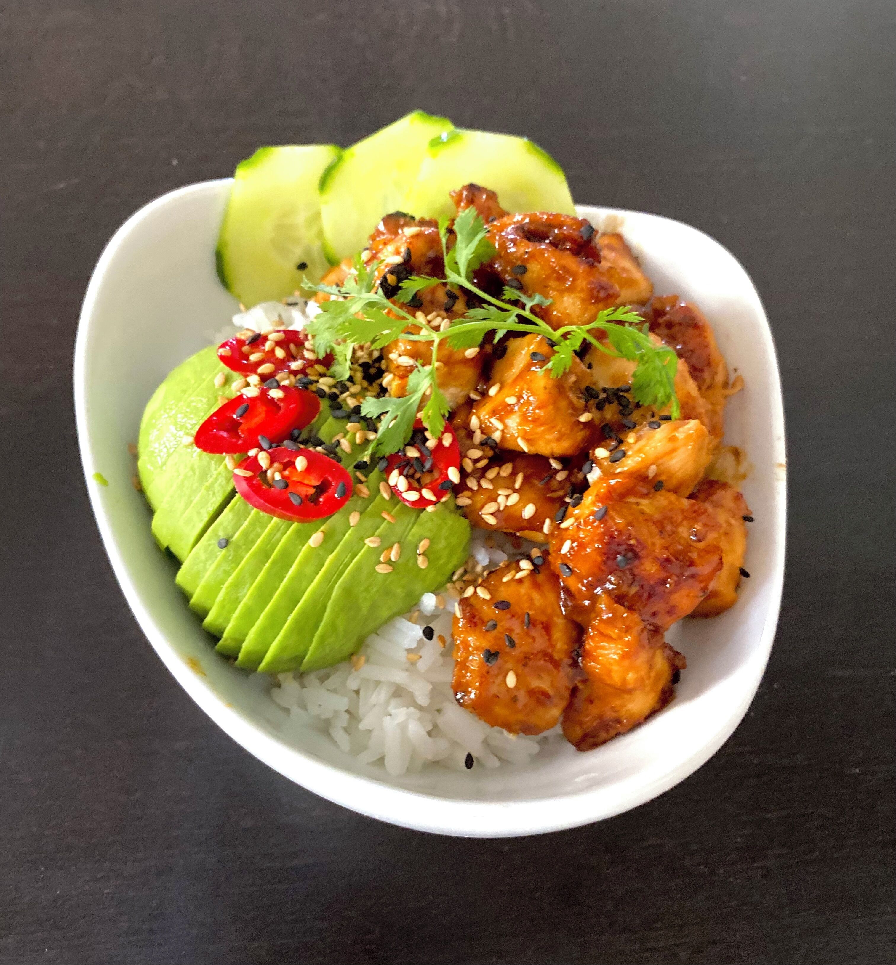 Un plato de comida con arroz, rebanadas de aguacate y un guiso de pollo decorado con semillas de sésamo y rodajas de chile rojo.