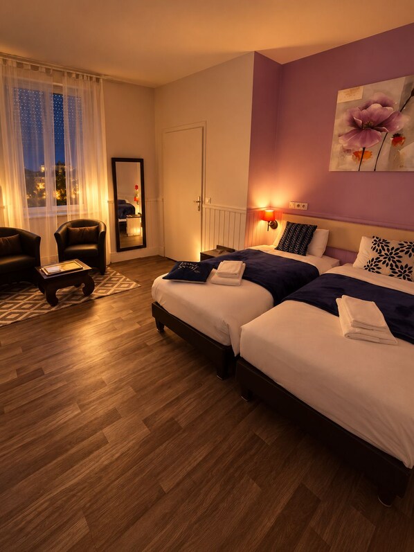 Habitación de hotel con dos camas, un área de descanso con una silla, un ventanal con cortinas y un cuadro en la pared.