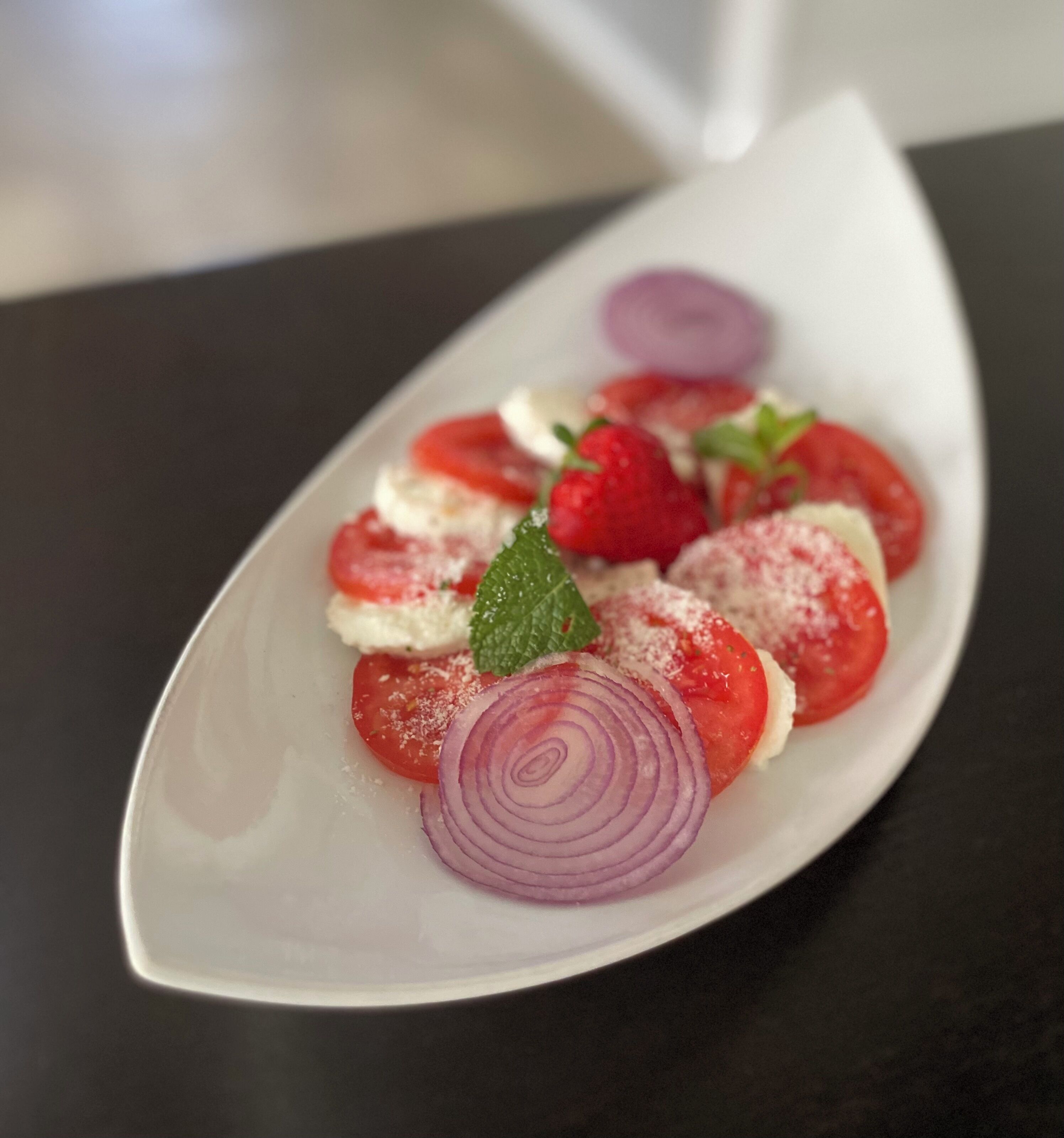 A white plate with a food arrangement including sliced tomatoes, a red onion, and a mint leaf.