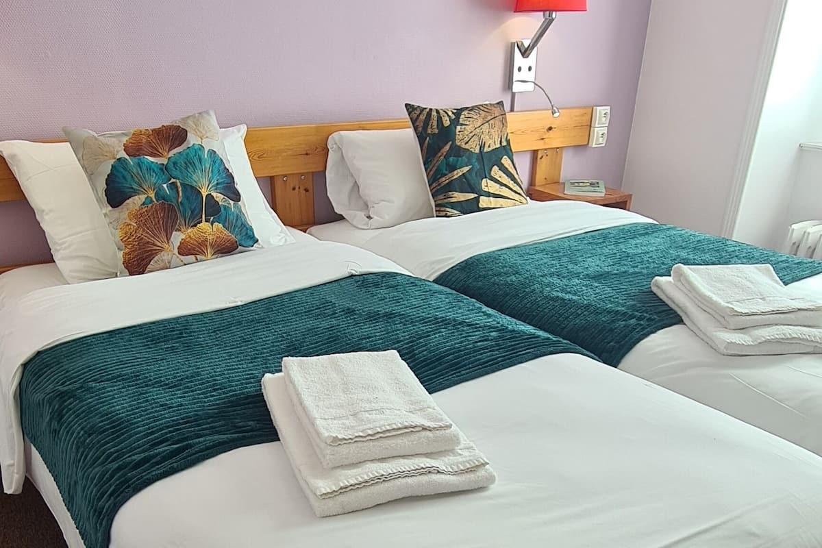 A hotel room with two beds, white linens, teal throw blankets, and a wall-mounted lamp.