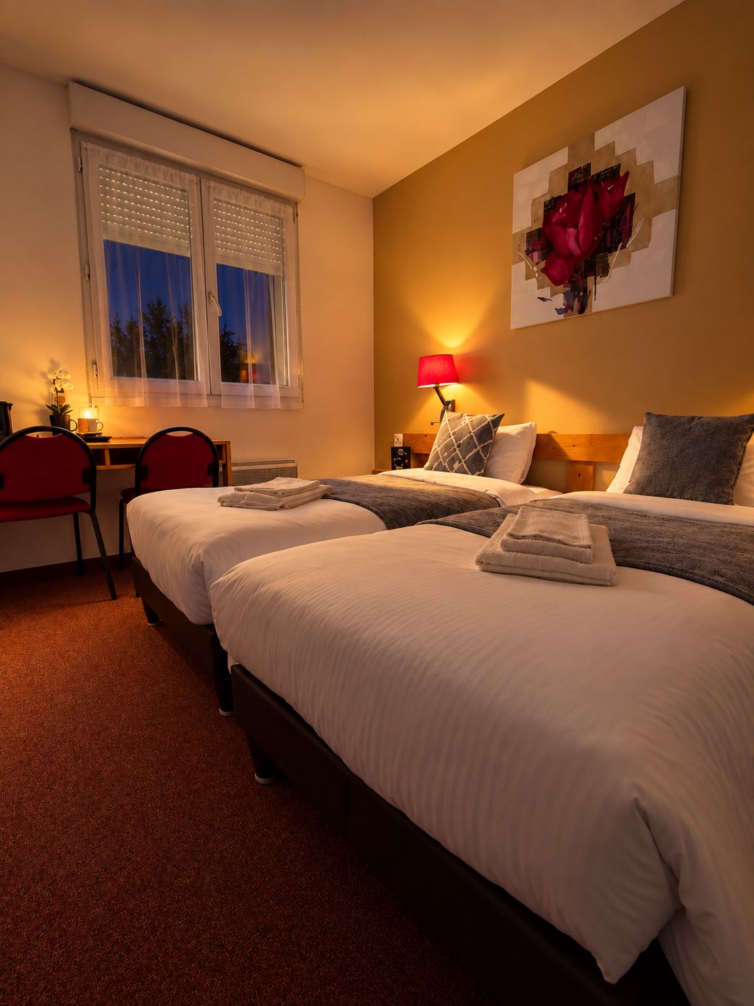 Een hotelkamer met twee bedden, een bureau met stoel, een lamp en een raam met jaloezieën.