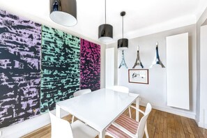 Uma sala de jantar moderna com uma mesa e cadeiras brancas, arte abstrata colorida na parede e luminárias pendentes.