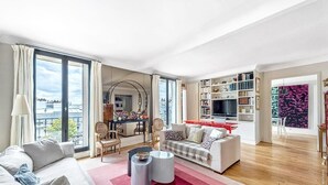Un saló modern amb un sofà, una taula de centre i una televisió. Hi ha una zona de menjador amb cadires i una taula. L'espai té grans finestres i vista a la ciutat.