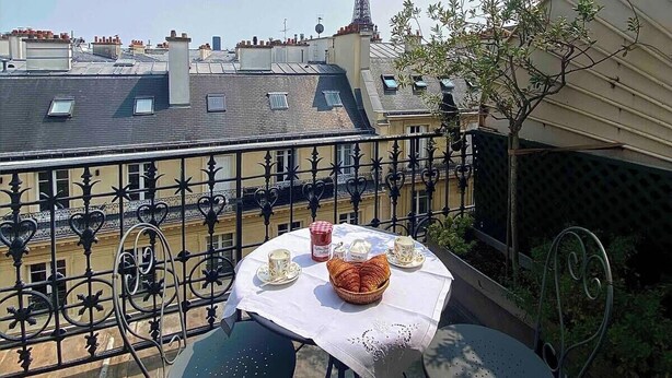 Un balcó amb taula preparada per esmorzar, amb croissants, tasses de cafè i una cistella. Es pot veure la Torre Eiffel al fons.