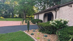 A house with a stone facade, a paved driveway, and a well-maintained garden with various shrubs and a flowering plant.