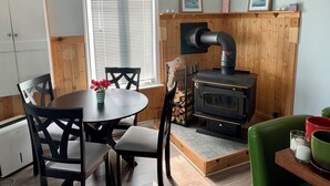 Een gezellige kamer met een houten kachel, een eettafel met stoelen, een groene fauteuil en een raam met jaloezieën.