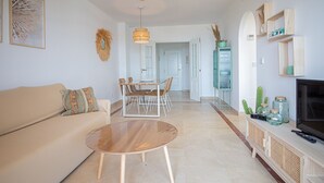 Uma sala de estar moderna com um sofá bege, uma mesa de centro redonda, um espaço de jantar com uma mesa branca e cadeiras de vime, uma TV sobre um suporte de madeira e um armário verde com potes.