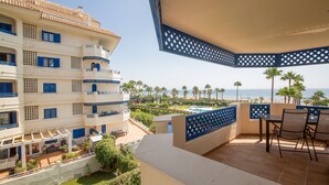 Varanda com vista para a praia e palmeiras, piscina e um edifício com persianas azuis e varandas.