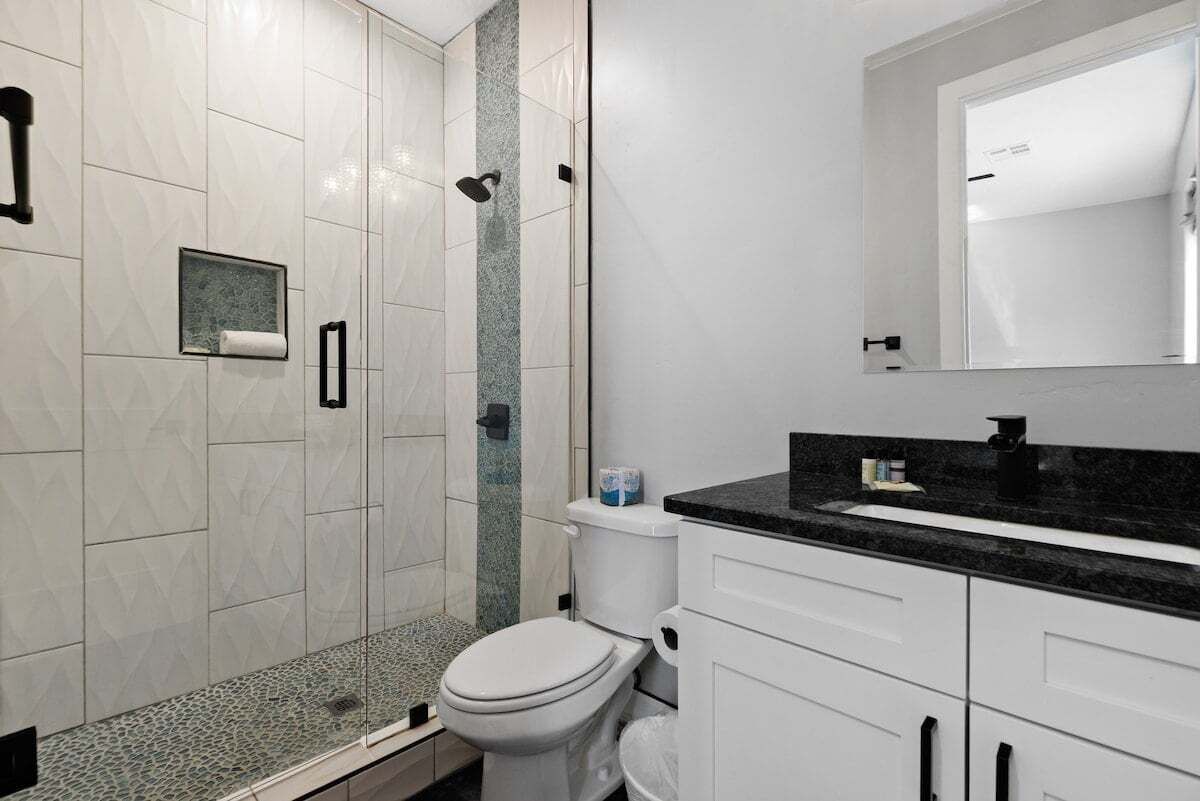 Une salle de bain moderne avec une douche à parois de verre, des carreaux blancs, un plan de toilette noir et des toilettes blanches.