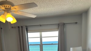 A bedroom with a bed, a window with a view of the ocean, a ceiling fan, and a nightstand with a lamp. - Ocean Front Condo (New Smyrna Beach)