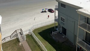 A beach view with a building in the foreground, a fenced area, and a few people on the sand. - Ocean Front Condo (New Smyrna Beach)