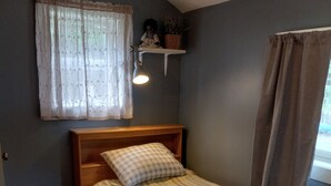 Un dormitorio con cama de madera, almohada de cuadros, colcha a rayas, ventana con cortinas traslúcidas, estante en la pared con una planta y una lámpara de noche.