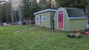 Un cobertizo de madera pequeño y verde con una puerta roja, ubicado en un área con césped y árboles al fondo.