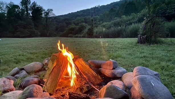 A campfire surrounded by rocks in a grassy field with trees and hills in the background.