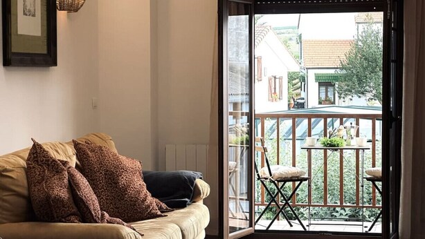 Una sala de estar con un sofá color beige, cojines estampados y un amplio ventanal corredera que da a un balcón con mobiliario exterior.