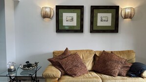 Una sala de estar con un sofá color beige, dos almohadones estampados, una mesa de centro de vidrio con un cuenco decorativo y dos lámparas fijadas a la pared.