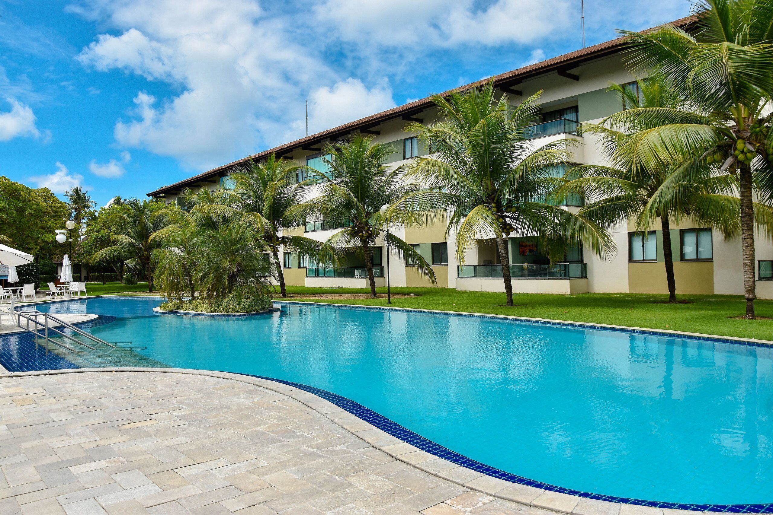 A swimming pool with a tiled edge, surrounded by palm trees and a building with multiple windows.