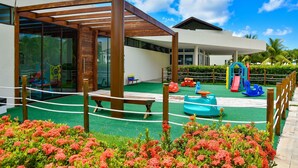 Un edificio moderno con un área exterior cubierta que incluye un banco de madera, una zona de juegos infantiles con toboganes y columpios, y un jardín con flores rosadas.