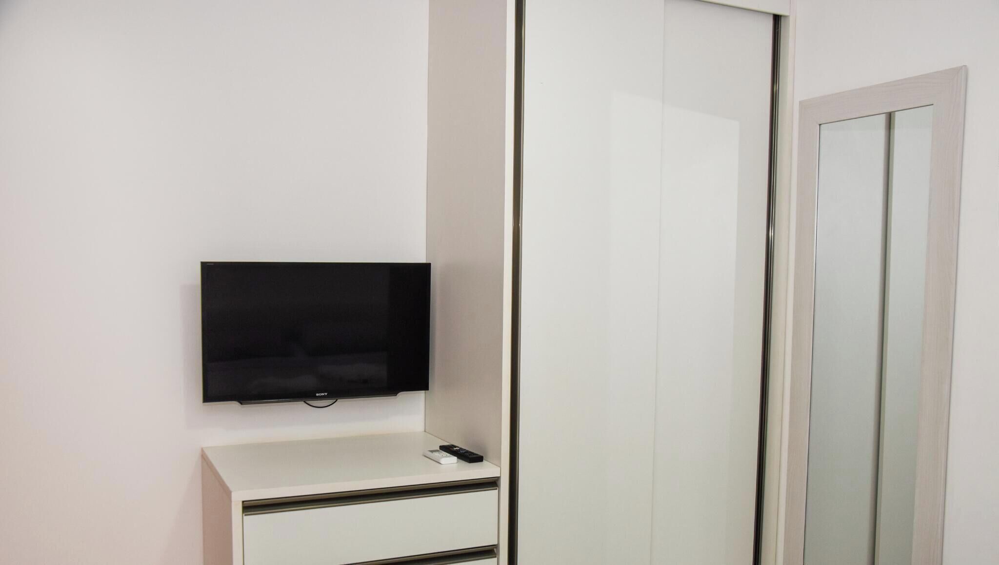 Un armario blanco con espejo, un televisor de pantalla plana montado encima y un pequeño escritorio blanco con un control remoto.