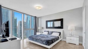 Een moderne slaapkamer met een groot bed, een bureau en uitzicht op hoge gebouwen.
