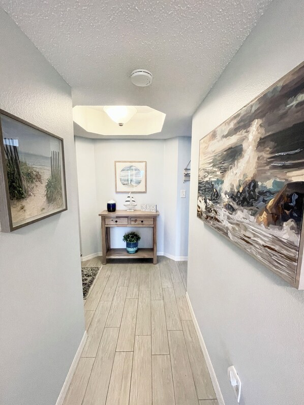 A hallway with a wooden floor, a ceiling light, a framed picture, a small table with a plant, and a large painting on the wall.