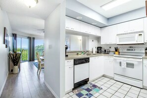Een moderne keuken met witte kasten, een vaatwasser, een magnetron en een oven. Er is een eethoek met een tafel en stoelen, en uitzicht op de omgeving door grote ramen.