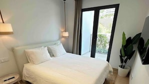 A modern bedroom with a large bed, a small side table, a potted plant, and a view of the outdoors through a sliding door. - Luxurious Flat With Jacuzzi and Pool at Higueron (Fuengirola)