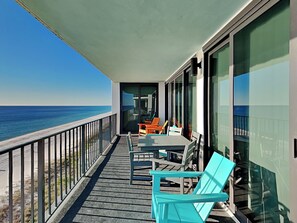 A balcony with a view of the ocean, a table with chairs, and a glass door leading to a room. - Watercrest #807 by Resort Collection (Panama City Beach)